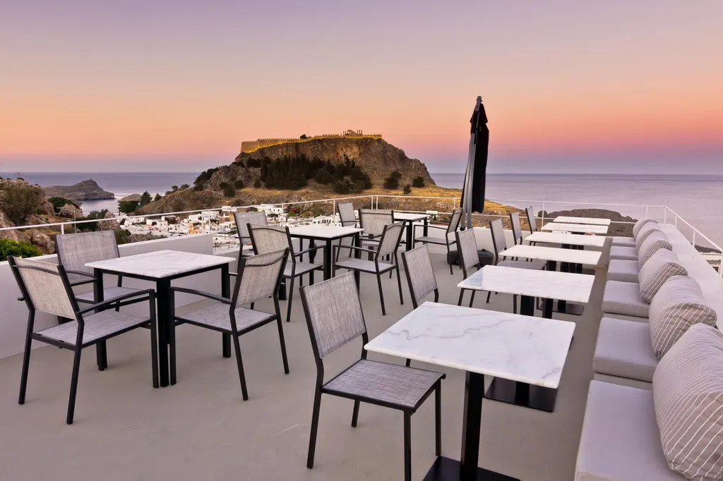 Terrace tables at Lindos Comfy Suites at dusk, with the Acropolis of Lindos under a pink sky
